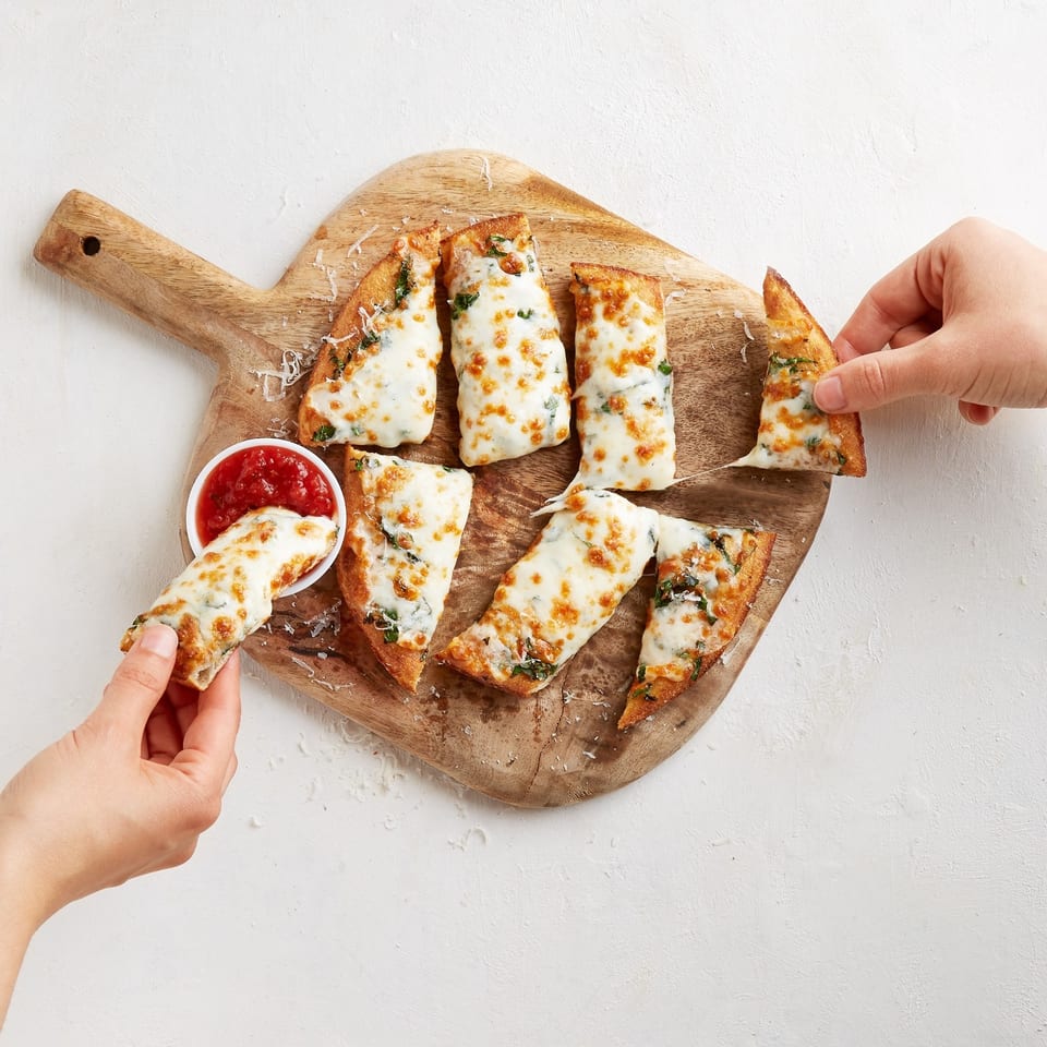 Hands grabbing cheesy garlic breadsticks with marinara sauce from a wooden board.