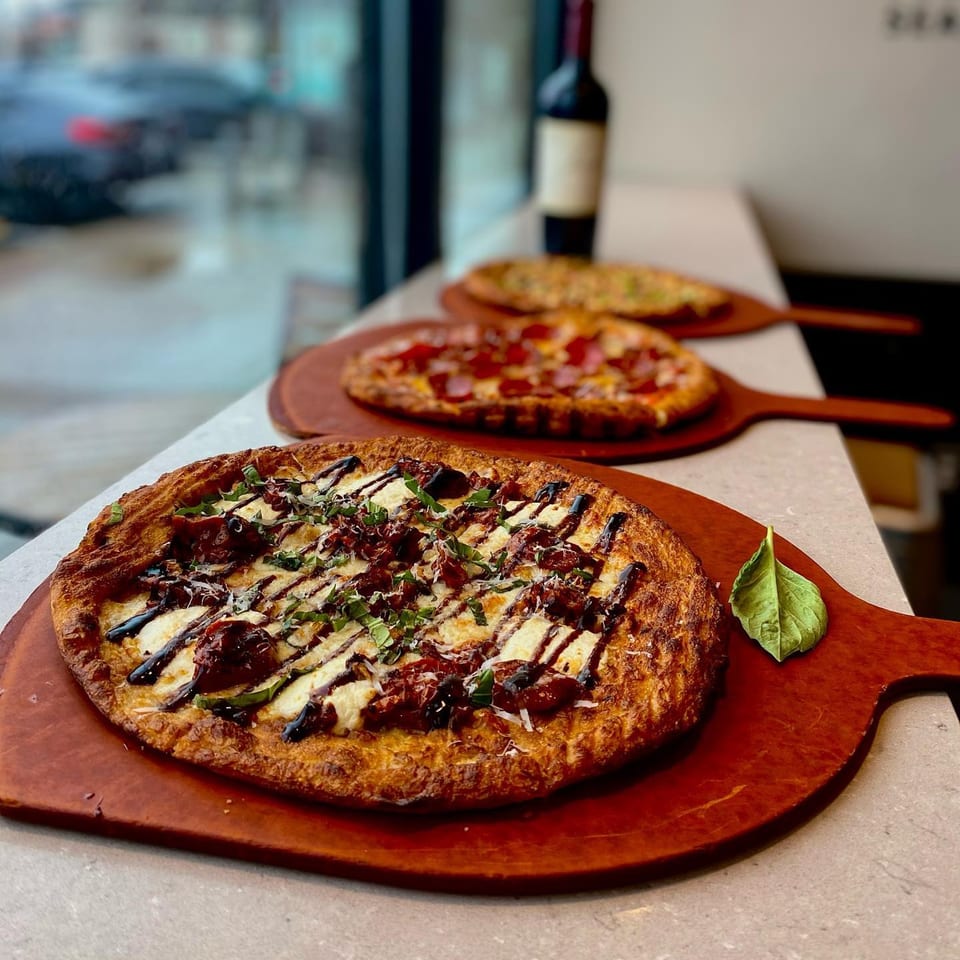 Three Oath pizzas on wooden peels, with a bottle of wine and a street view in the background.