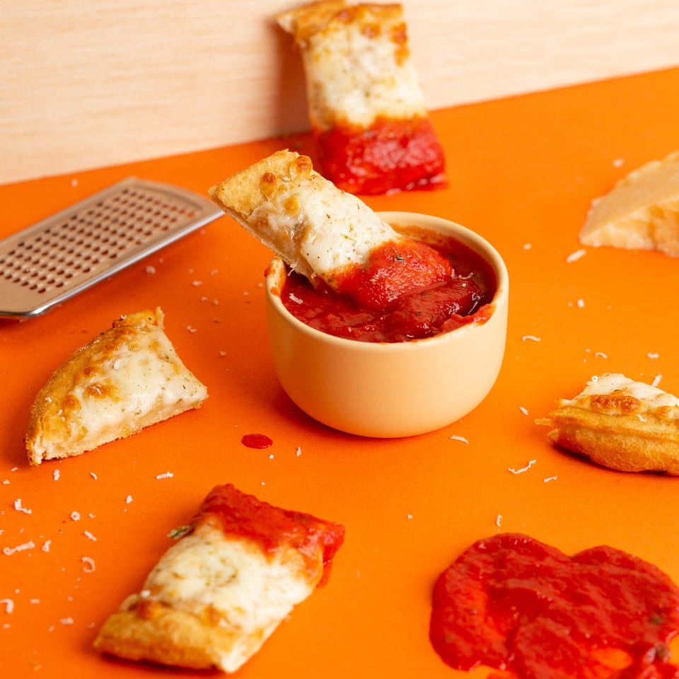 Oath Pizza breadsticks with melted cheese, dipped in marinara sauce, on an orange background.