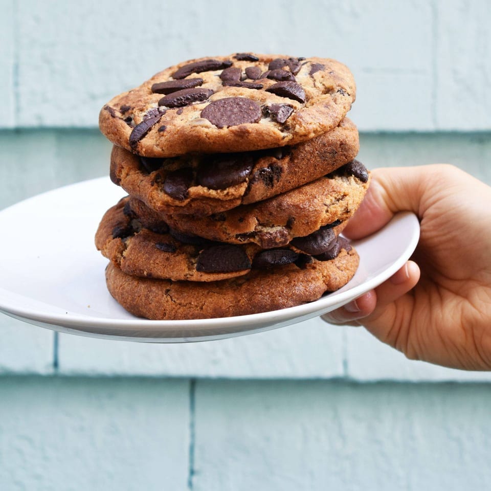 A hand holds a white plate with a tall stack of Oath Pizza chocolate chip cookies.